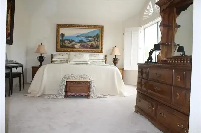 Bedroom with large bed, decorative cover, landscape painting, wooden furniture, and windows