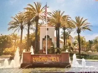 Sign reading "MARINA HILLS" with building, palm trees, fountains, and American flag