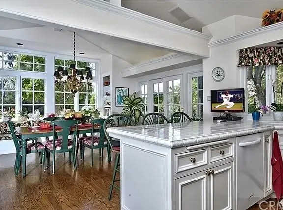 Kitchen with white cabinets, large island, hardwood floors, green dining table and chairs, TV, and plants