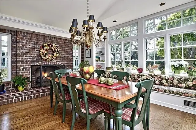 Dining area with wooden table, green chairs, chandelier, brick fireplace, windows, and window seat with cushions