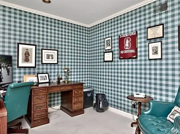 Room with green and white wallpaper, wooden desk, framed pictures, trash bin, teal chair, and Stanford banner