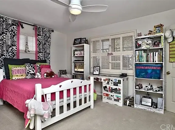 Bedroom with pink bedspread, decorative pillows, white bed frame, window with curtains, bookshelves, carpet, and ceiling fan