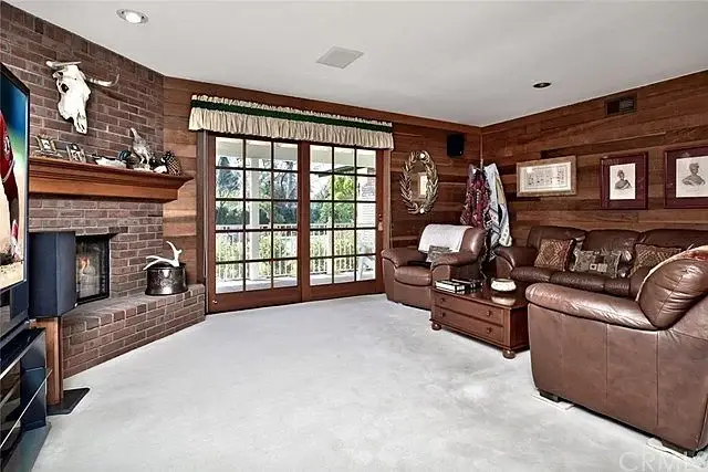 Living room with brick fireplace, wooden paneling, leather couches, windows, and decorations