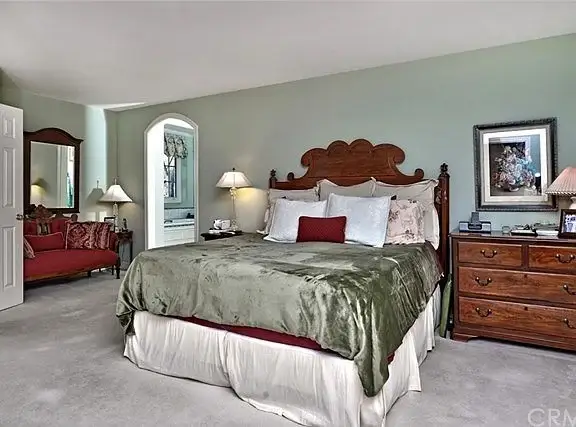 Bedroom with large bed, green bedding, wooden headboard, pillows, lamps, dresser, and armchair