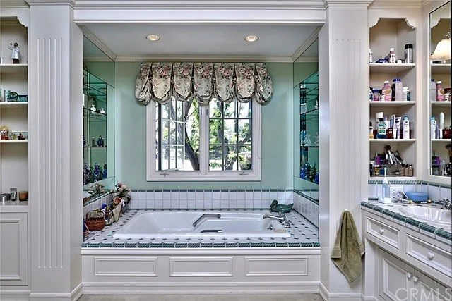 Bathroom with large bathtub, white cabinets, shelves, and window with curtains