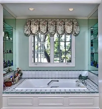 Bathroom with large bathtub, tiled surround, plants, window with floral curtains, and glass shelves
