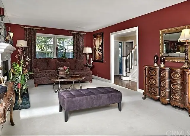 Living room with maroon wall, dark sofa, coffee table, ottoman, wooden cabinet, lamps, and stairs
