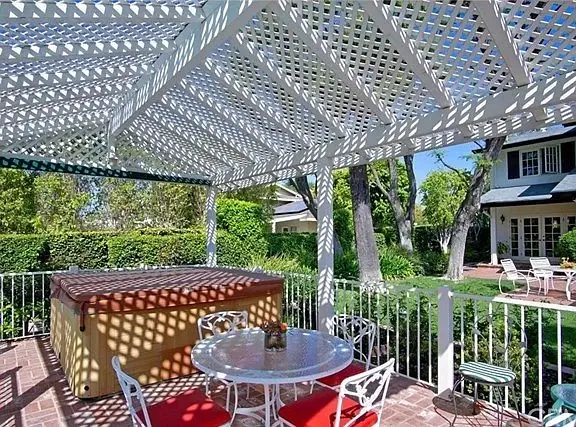 Patio with white lattice roof, hot tub, table with red cushions, greenery, and house California real estate in background