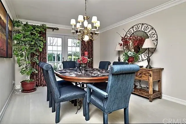 Dining room with round wooden table, upholstered chairs, chandelier, plants, artwork, and sliding glass doors