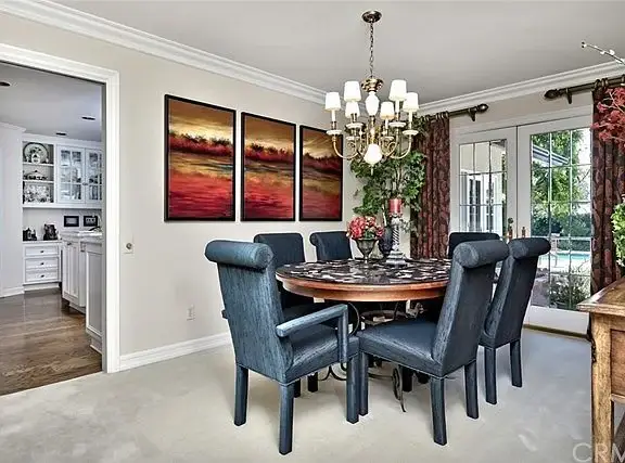 Dining room with round table, six blue chairs, chandelier, paintings, and windows