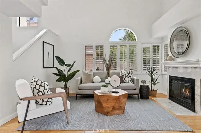 Living room with gray sofa, coffee table, armchair, fireplace with round mirror, plants, and large windows