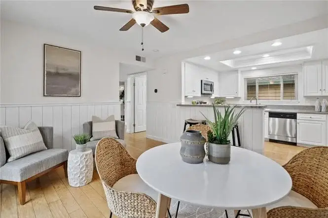Dining area with round white table, woven chairs, wooden floor, beige furniture, and kitchen with white cabinets and stainless steel appliances