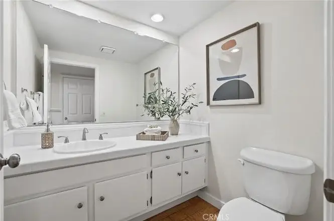 Bathroom with double sink vanity, white cabinets, framed artwork, towel rack, toilet, and wood floor