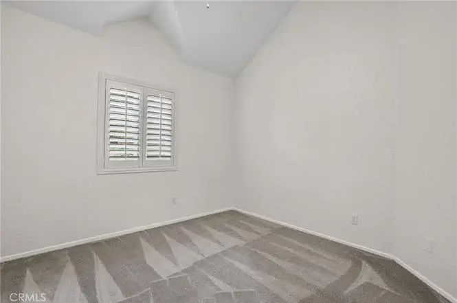 Empty room with vaulted ceiling, white walls, carpet, and window with shutters