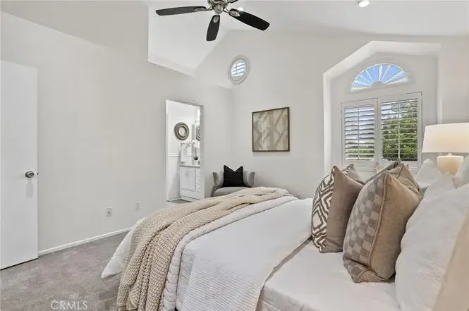 Bedroom with made bed, decorative pillows, ceiling fan, windows with shutters, mirror, and carpeted floor