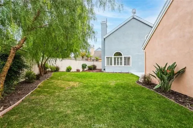 Backyard with green grass, shrubs, trees, pale blue house with chimney, and fenced