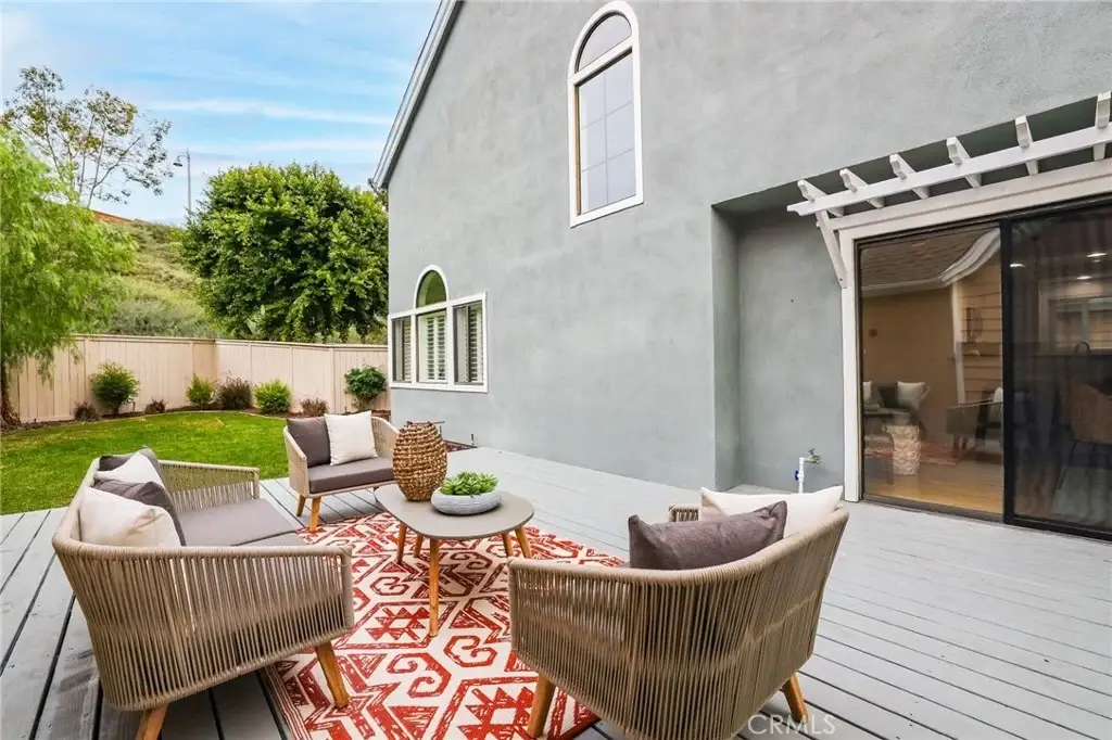 Outdoor patio with wicker chairs, round table, patterned rug, gray house exterior, and greenery