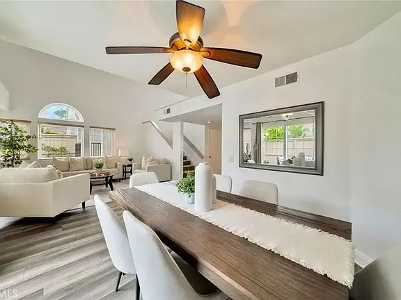 Dining area with wooden table, white chairs, ceiling fan, big windows, and mirror on the wall.