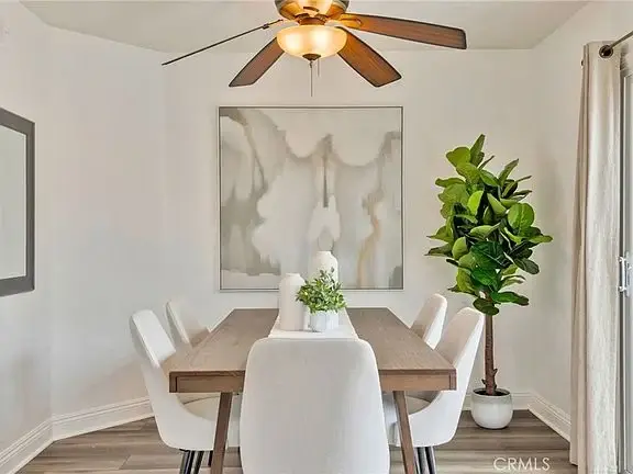 Dining area with wooden table, white chairs, ceiling fan, plant, and wall art.