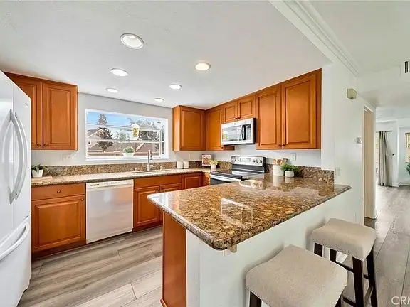 Kitchen with wooden cabinets, granite counter, stainless appliances, sink window view, and bar stools.