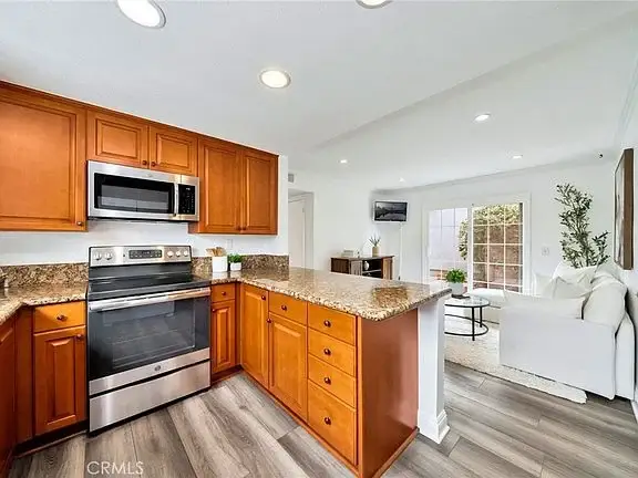 Kitchen with wooden cabinets, stainless appliances, granite counter, oven, microwave, and fridge with living room in back.