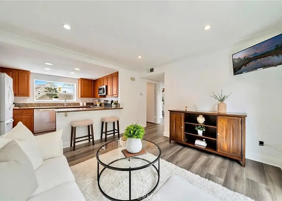 Living room and kitchen with wooden cabinets, bar stools, sofa, coffee table, TV, and shelves with decor.