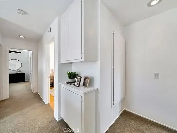 Hallway with beige carpet, white walls, lights, small cabinet with photos, and plant.