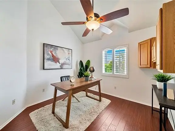 home office with wooden desk, black chair, plants, wall art, ceiling fan, and window with shutters.