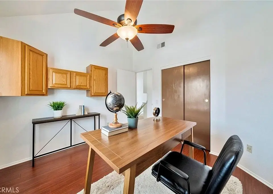 Home office with wooden desk, globe, books, plant, black chair, wall cabinets, and ceiling fan.