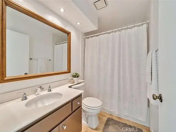 Bathroom with large mirror, sink with three faucets, shower curtain, toilet, and shelf decor.