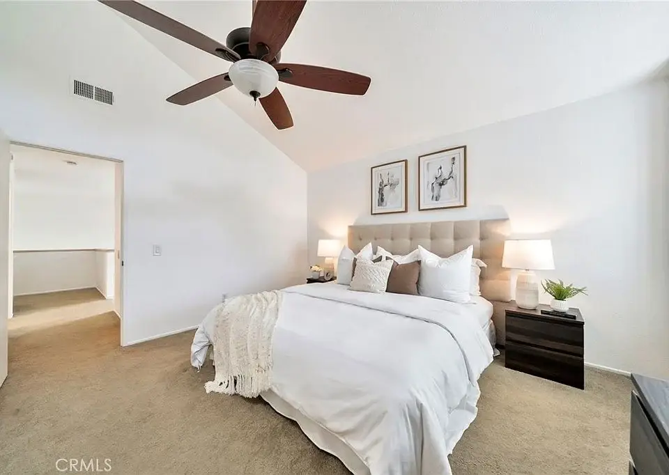 Bedroom with white bedding, lamps, wall art, ceiling fan, and light carpet.