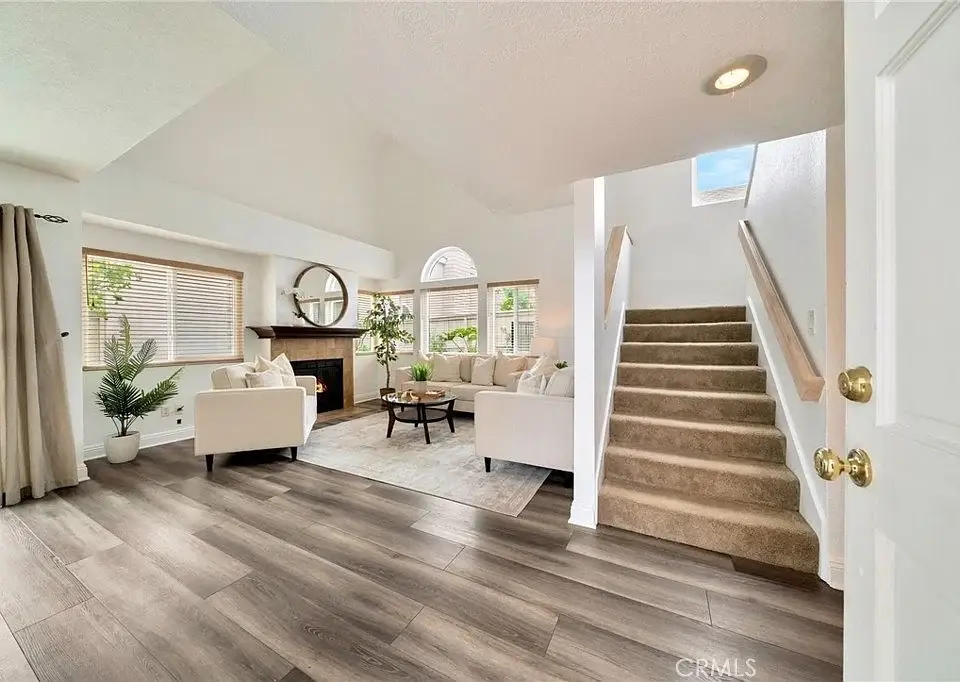 Living room with stairs, big windows, round mirror, plants, and light furniture.