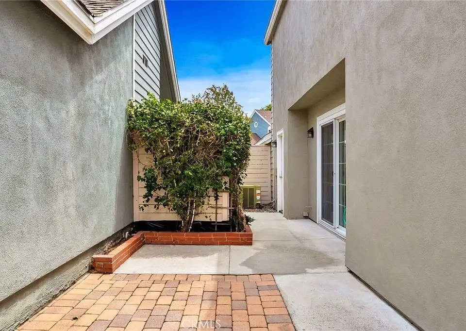Pathway between two buildings with plants on one side and a paved area in front.