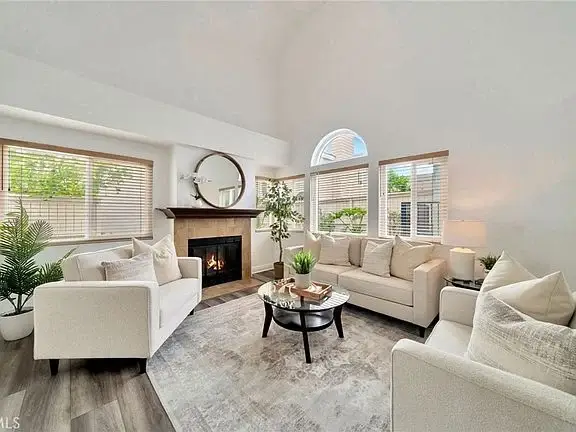 Living room with two white sofas, round coffee table, gray rug, fireplace, big windows, and plants.