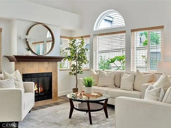 living room with white couch, pillows, round coffee table, stone fireplace, big windows with blinds, and a plant.