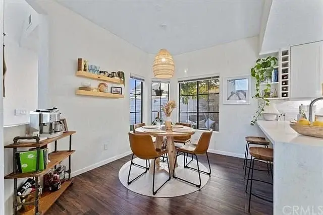 Dining area with round table, four chairs, wall shelf, plants, and kitchen counter visible