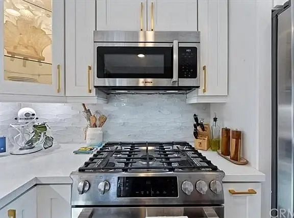Kitchen with gas stove, stainless steel microwave, white cabinets, marble backsplash, and countertop utensils