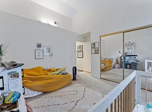 Bedroom with bed, yellow blanket, white rug, mirrored closet doors, and shelf decor