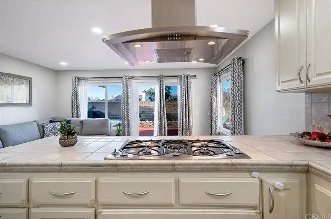 Kitchen with gas stove, windows, and living area with gray couch and patterned curtains