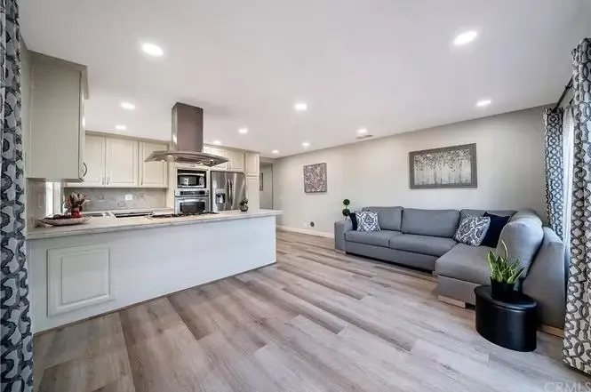Living room and kitchen with gray sofa, wooden floors, large window, and stainless steel appliances