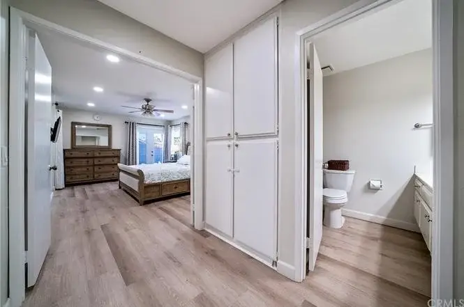 Hallway with wooden floor and neutral walls, leading to bedroom with bed and dresser, and bathroom with toilet