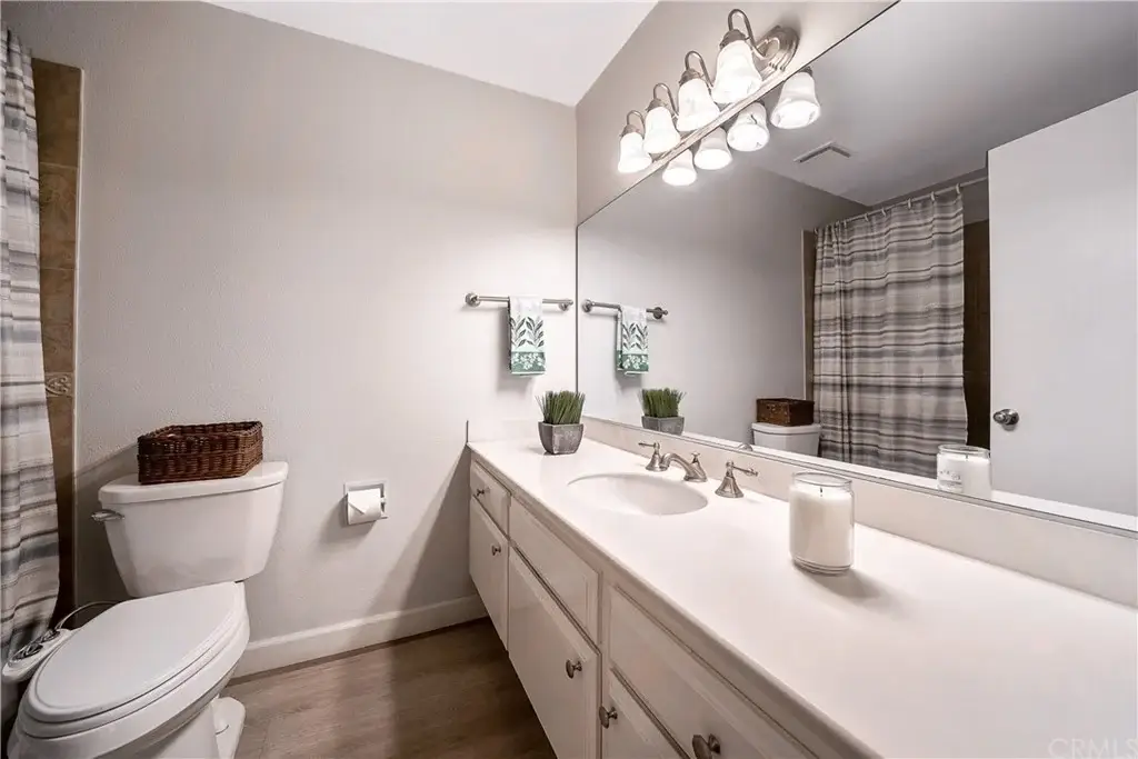 Bathroom with white dual-sink vanity, large mirror, beige walls, shower curtain, toilet, and decorative plants