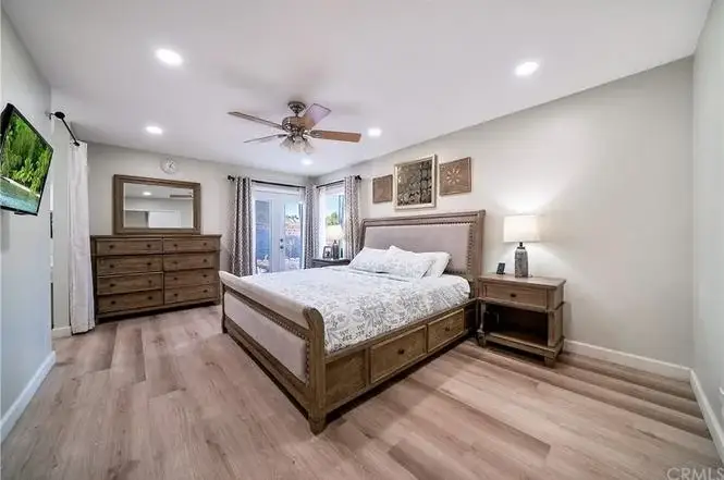 Bedroom with queen bed, light patterned comforter, dresser with mirror, nightstand with lamp, TV, and large windows