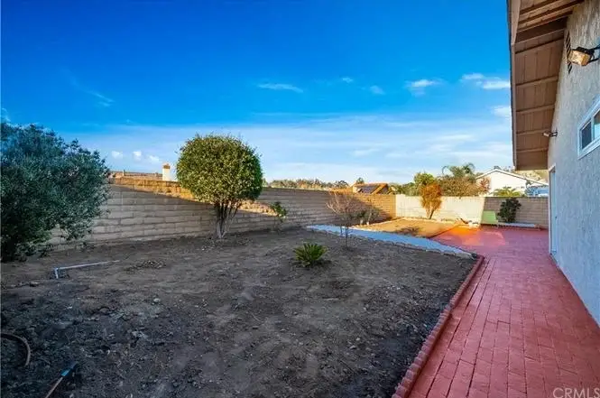 Backyard with dry soil, small plants, paved walkway, and brick pathway