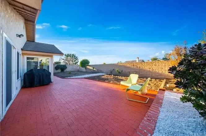 Backyard patio with red brick floor, grill, two chairs, plants, and wall in background