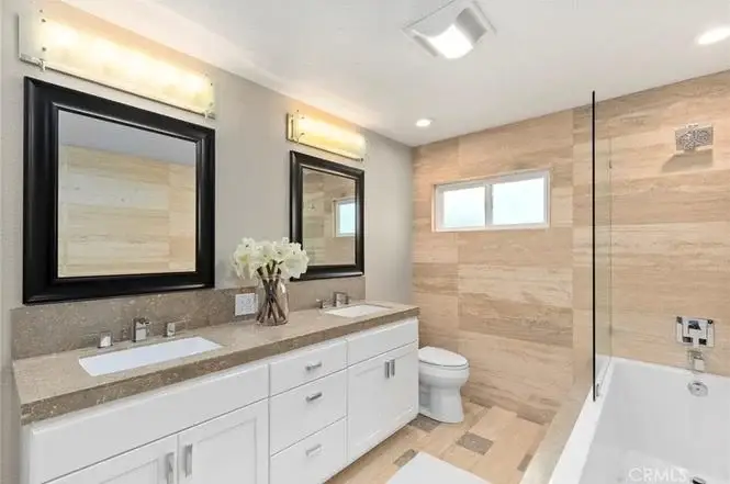 Bathroom with double sink vanity, large mirrors, stone walls, glass tub, toilet, and flowers.
