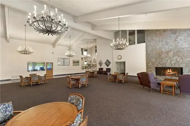 Large room with high ceiling, chandeliers, stone fireplace, tables, chairs, and big windows.