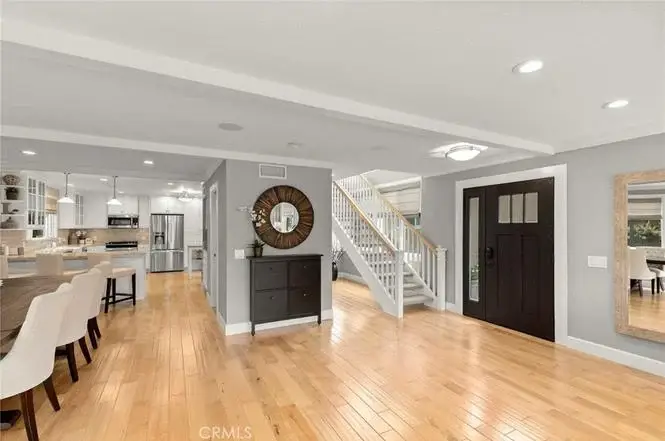 Open living space with wood floor, dark front door, stairs, kitchen with white cabinets, bar seats, and wall mirror.
