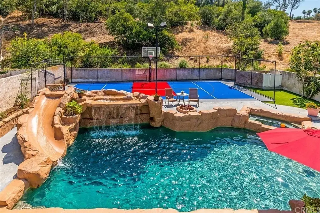 Backyard of house California real estate with pool, water slide, stone structure, basketball court, chair, umbrella, and plants.