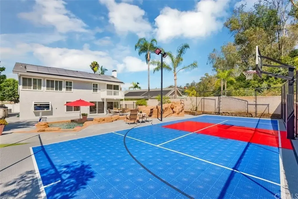 Outdoor basketball court with blue and red surface, greenery, and large luxury house California real estate in background